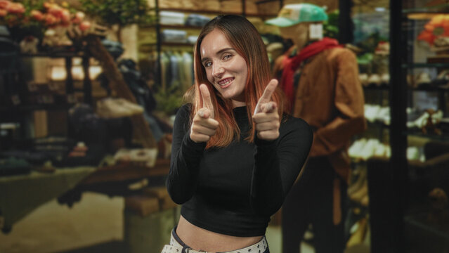 Woman smiling and making finger guns gesture toward camera among mannequins, racks and a jacket in a boutique building; playful confidence.