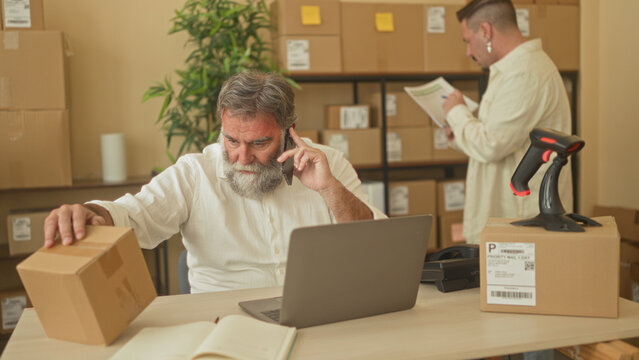 Man on phone types at laptop while handling a parcel and reaching across desk in a small business building; teamwork efficiency concentration.