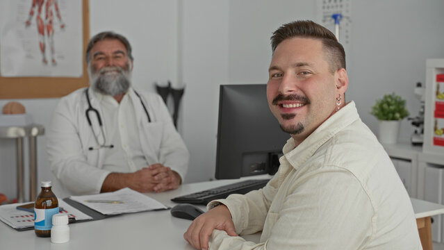 Two men, a doctor wearing a stethoscope and a smiling male patient with hands resting on a desk, pill bottles visible, seated in a clinic building; trust.