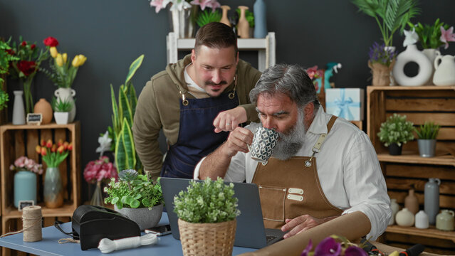 Two men florists in aprons lean over a laptop, one points finger to screen and gestures at online order while the older bearded florist holds a mug in a flower studio; focused teamwork.