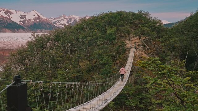 Woman walking across a suspended bridge surrounded by the natural beauty of forests, glaciers, and mountains in the distance, Torres del Paine, Patagonia, Chile