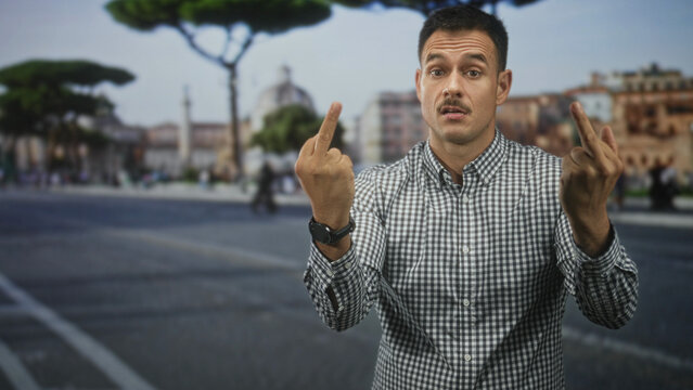 Man with moustache and checked shirt shows both middle fingers in street square with stone pavement and umbrella pine trees, confrontational gaze; anger defiance.