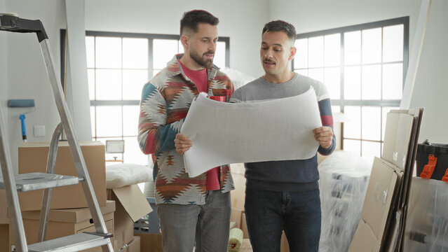 Man holding blueprint with both hands and reading plan in an apartment full of boxes and ladder; home planning anticipation.
