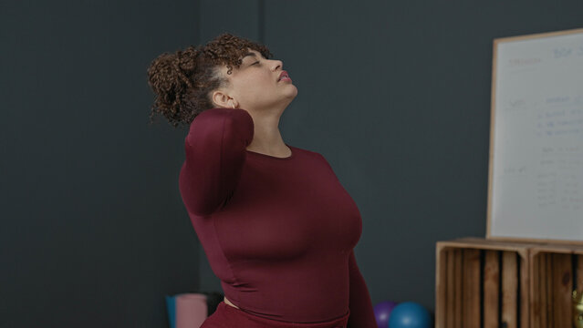 Woman massaging neck and stretching shoulder in studio with yoga mat and exercise balls; relief recovery wellness.