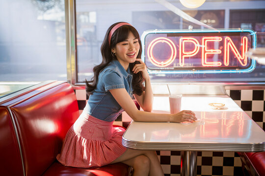 Young woman smiles warmly at diner booth. Neon 'OPEN' sign glows behind her. Red vinyl seat and checkered floor add retro vibe. Perfect for advertising, food service, or vintage themed content