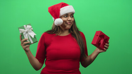 Fototapeta premium Woman holding two gift boxes, hands visible, wearing santa hat and red top against green studio backdrop; cheerful celebration.