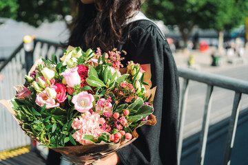 Fototapeta premium A side profile of a university graduate wearing a black academic gown and holding a vibrant flower bouquet filled with pink roses, carnations, and lush green foliage, positioned next to a metal bridge