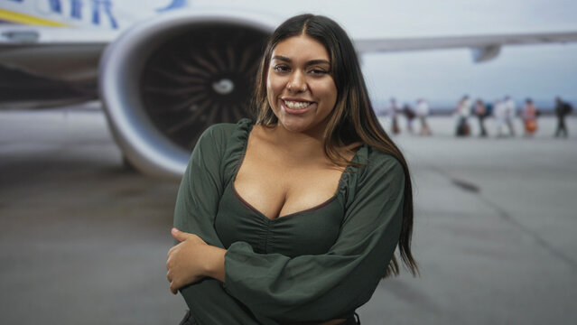 Woman smiling with arms crossed over cleavage in green blouse beside airplane engine on tarmac at airport with passengers boarding in background; confidence.