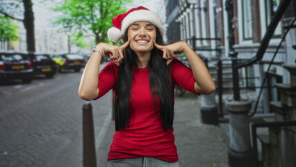 Fototapeta premium Young woman in santa hat presses cheeks with index fingers, smiling broadly on a city street; holiday cheer joyful.