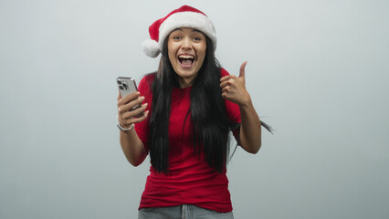 Fototapeta premium Young woman in santa hat holding smartphone and showing thumbs up in studio, wearing red shirt; joyful holiday celebration.