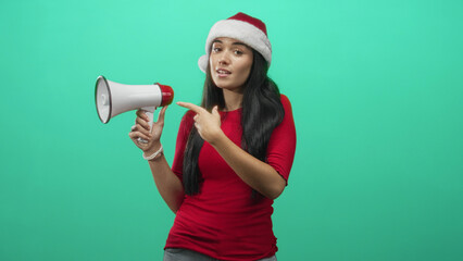 Fototapeta premium Woman in santa hat pointing finger at megaphone in studio, red top and exposed hand gesture for announcement; joyful holiday spirit.