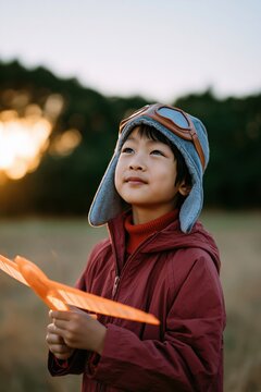 Child with orange toy plane looking toward the sky at sunset in a field wearing an aviator cap for kid banner and outdoor background design