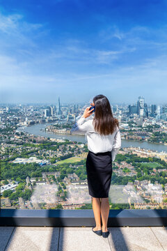 A business woman talking on her phone and looking at the elevated view of the skyline of London, England, through large office windows