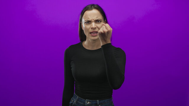 Woman in black long sleeve top and jeans beckons with curled hand and furrowed brow toward camera in studio with purple backdrop; anger defiance.