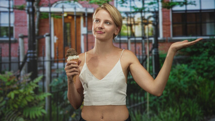 Fototapeta premium Blond woman holding a double scoop ice cream cone and gesturing palm up while standing before a brick building and iron fence; curiosity leisure summer.