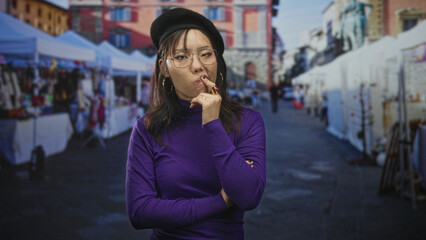 Fototapeta premium Woman touching finger to lips on a street lined with market stalls, wearing purple turtleneck sweater, beret and glasses; pensive curiosity.