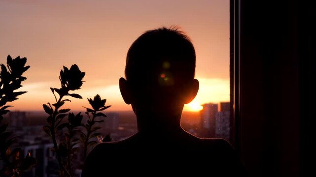 Dark silhouette of child head from behind watching sunset over city. A lonely boy looking out window at beautiful orange sky, dreaming and thinking about the future. View from high rise house
