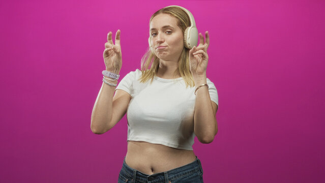 Woman wearing over ear headphones holds ear cup and makes peace sign with air quotes, showing exposed belly against pink studio backdrop; playful confidence.