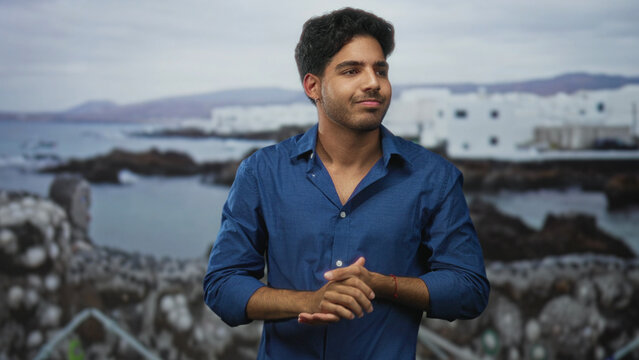 Man in blue shirt clasping hands and showing forearms in studio with a seaside backdrop of white buildings and rocky shore; calm introspection.