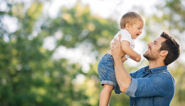 Father lifts and plays with his happy toddler son outdoors against a blurred green background. The joyful interaction highlights family bonding and parenting moments.