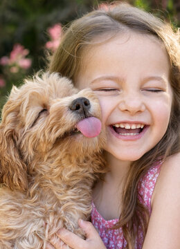 Young girl embraces a fluffy puppy while laughing with eyes closed in a sunny garden. The moment captures joy and affectionate play between child and dog.