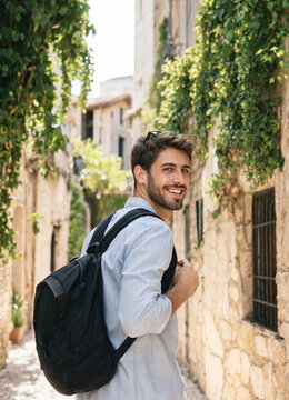 Young man walking with a backpack and smiling, looking back over his shoulder in a sunlit stone alley lined with greenery. Casual travel mood and warm natural light.