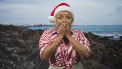 Fototapeta premium Senior woman wearing red santa hat, glasses and striped shirt claps hands, covers mouth, then opens palms in studio with rocky shore backdrop; holiday surprise joy.