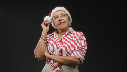 Fototapeta premium Woman in red striped shirt wearing glasses touches santa hat pompom to her ear while arms are crossed in studio; nostalgic holiday reflection.