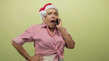 Fototapeta premium Woman, senior, wearing red santa hat and red striped shirt, holding smartphone to ear with hand on hip in studio; holiday surprise.
