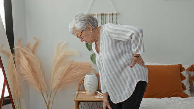 Woman holding lower back and bending beside bed in a bedroom of a residential building; discomfort aging.