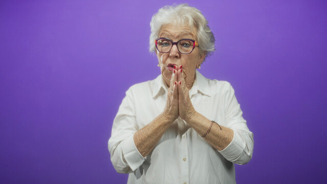 Woman elderly with white hair and eyeglasses, palms together prayer gesture, closed eyes in a purple studio set wearing white shirt and bracelet; serenity reflection.