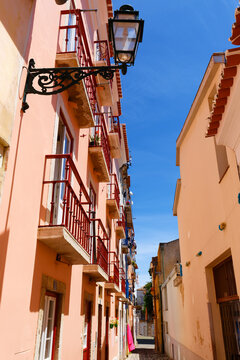 Charming narrow Lisbon street bathed in warm sunlight, lined with a long pink-rendered building featuring stacked red-painted iron balconies with potted flowers.