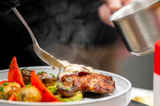 Chef pouring creamy mushroom sauce over grilled chicken steak with roasted vegetables.