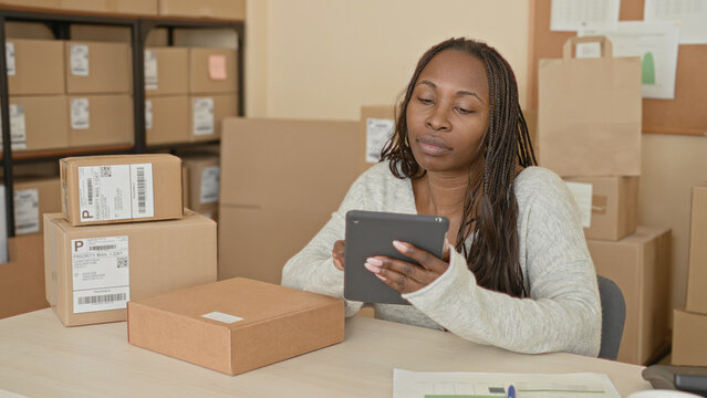 Woman holding tablet among stacked shipping boxes in a building workspace checking parcels and labels with a thoughtful expression; small business optimism.