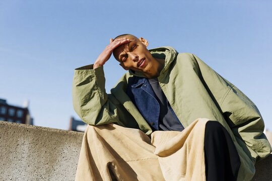 Fashion portrait of a person in an oversized green jacket and beige wide pants sitting on a concrete rooftop ledge for editorial streetwear background