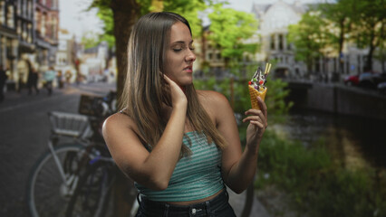 Fototapeta premium Woman holding an ice cream cone, hand to neck while inspecting dessert on a busy cobblestone street beside a canal with bicycles and trees; hesitation.