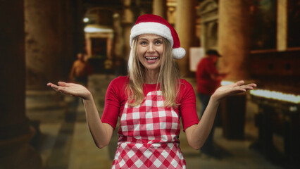 Fototapeta premium Woman wearing santa hat and red checkered apron shows open palms and smiles while shrugging inside a dim building aisle with candles and pillars; holiday cheer.