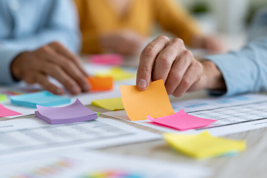 Hands arrange colorful sticky notes on documents. People collaborate, focused, creative. Ideal for brainstorming, project planning, team meetings. Soft lighting adds calm, productive atmosphere