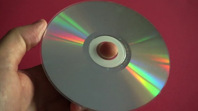 Shiny compact disc held in a hand reflecting rainbow colors on its surface against a red background