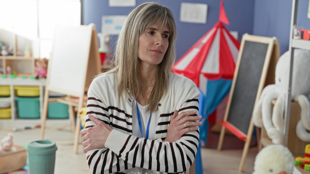 Woman teacher with arms crossed, lanyard visible and coffee cup on table in a preschool classroom; quiet concern.