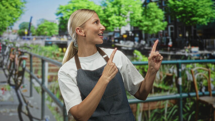 Fototapeta premium Blonde woman in apron points finger with both hands toward a canal railing on a busy street in daytime; playful surprise.