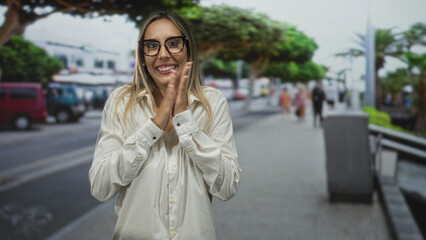 Fototapeta premium Woman clapping hands on city street sidewalk, smiling with glasses and white shirt while parked vans and trees blur behind her; joy.