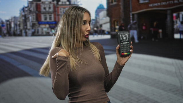 Woman points finger to card reader while holding payment terminal on a busy city street near shops; payment confusion.