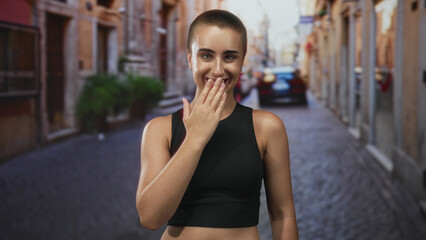 Fototapeta premium Young woman with short hair covers mouth with hand while blowing a kiss on a narrow cobblestone street in a city alley; playful.