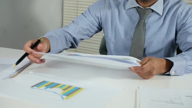 A young businessman is sorting through files scattered on his desk. A busy workspace is visible. Documents and files are visible, highlighting the organization, record-keeping, and daily administrativ