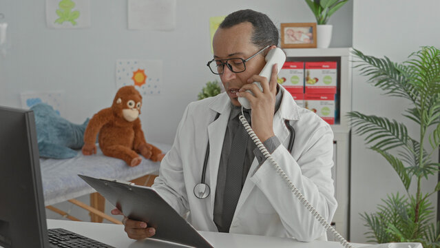 Man doctor with stethoscope holding telephone to ear while checking charts at clinic; focused professional care.