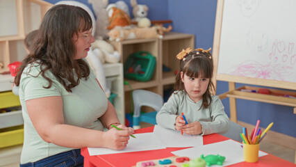 Naklejka premium Woman holds marker guiding girl drawing at table with paper and colored pencils in kindergarten classroom building; learning joy.