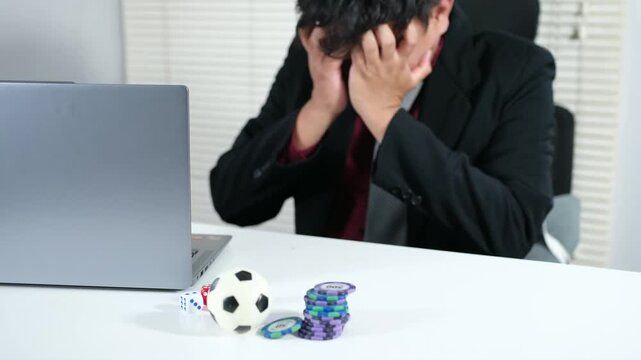 A stressed man loses in online soccer gambling with dice, a soccer ball, and a laptop on the table. A gambler loses a lot of money playing soccer, a gambling addiction. Loss, ruin, debt, the concept o