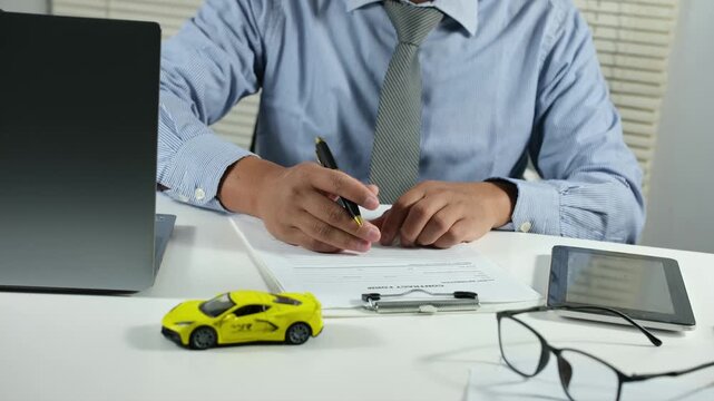 A man buys or sells a car. A businessperson signs purchase documents, car insurance, or a lease. Someone signs a contract or agreement. A yellow toy car sits on a table, selective focus.