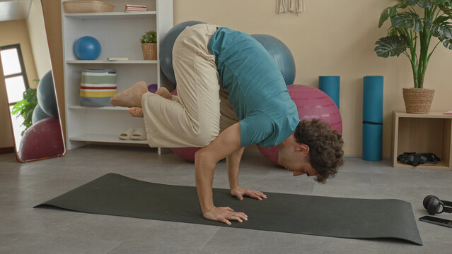 Young hispanic man holds crow pose on yoga mat using hands for balance in a studio surrounded by exercise balls and foam rollers; calm focus.
